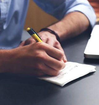 Homem sentado em uma mesa, escrevendo em um caderno enquanto olha o notebook [foco apenas em seus braços]