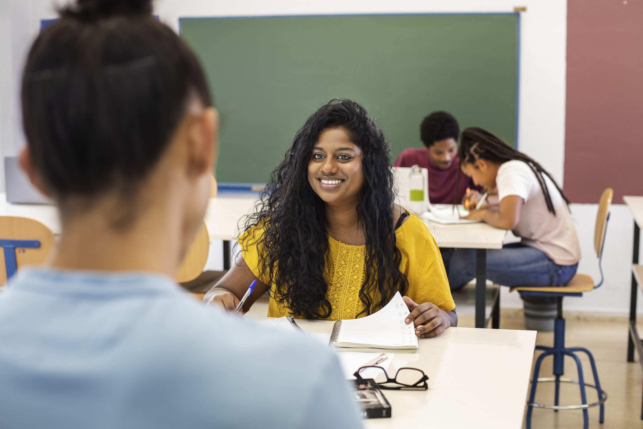 Estudante sorri para professora a sua frente, enquanto colegas ao fundo fazem tarefas na sala de aula