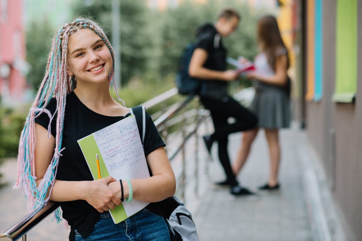 estudante universitária posa sorridente no campus segurando um caderno nos braços com colegas ao fundo enquanto se preparam para o Vestibular PUC Campinas 2024