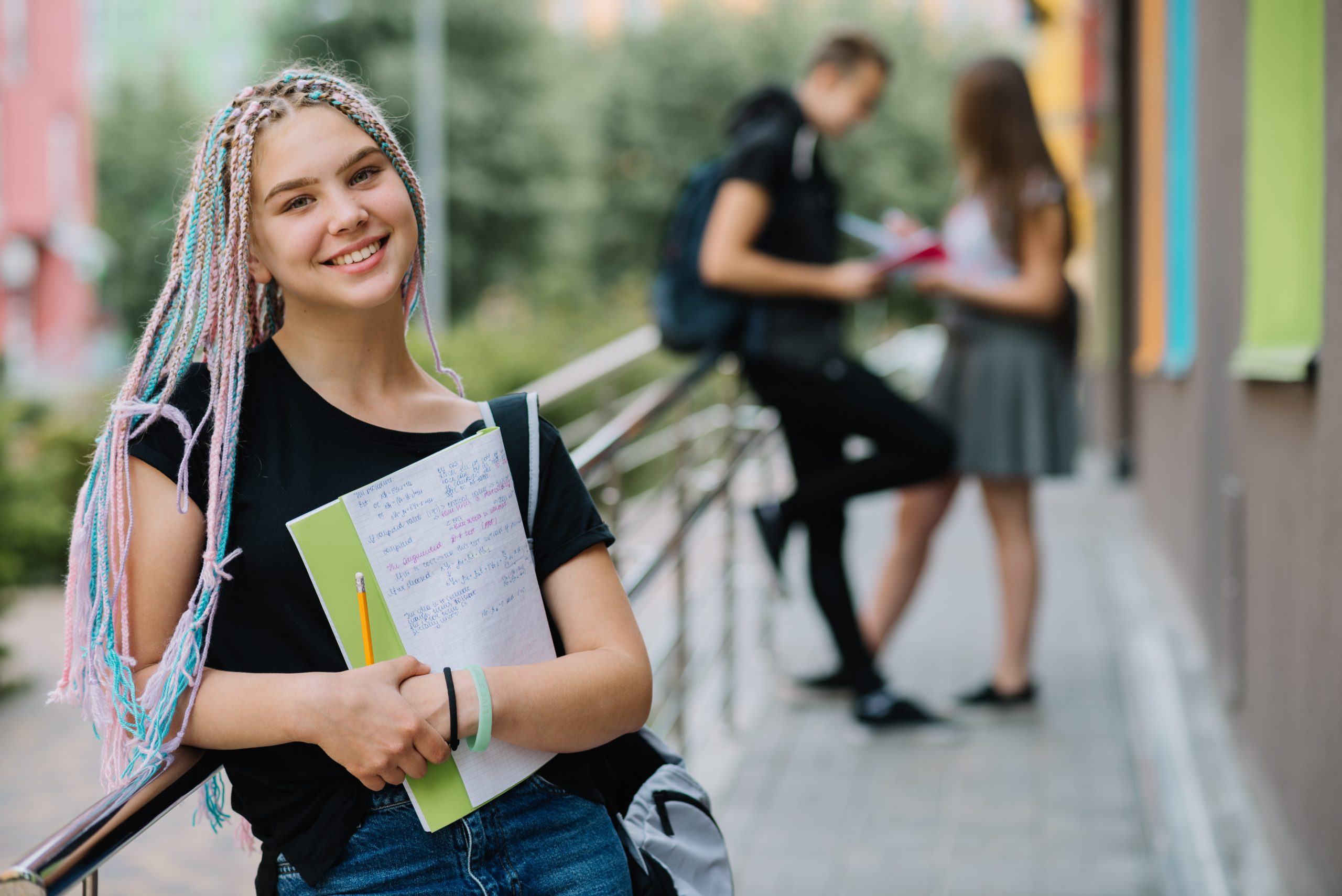 Estudante universitária posa sorridente no campus segurando um caderno nos braços com colegas ao fundo