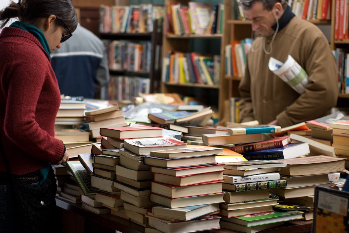 Duas pessoas aparecem em um sebo, há inúmeros livros empilhados em cima de uma mesa, onde ambos olham as opções disponíveis 