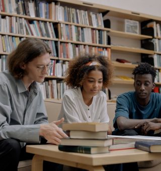 Três alunos estudando para o Vestibular Filosofia 2024. Eles estão sentados em um frete a alguns livros dentro da biblioteca