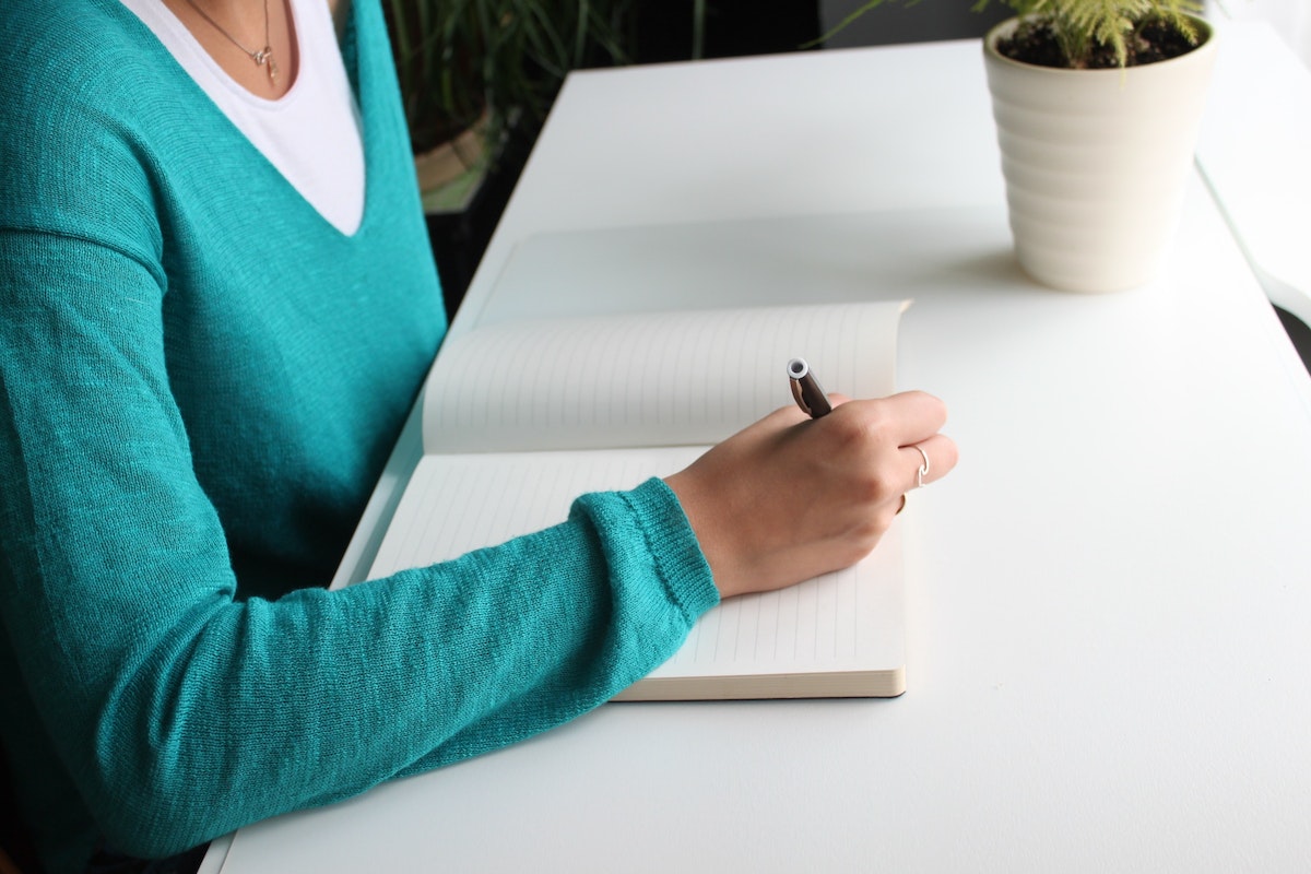 Uma mulher escrevendo em um caderno branco que esta em cima da mesa. Ela anota a lista de Universidades sem Vestibular em 2024