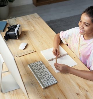 Uma menina sentada na frente do computador com seu caderno na frente enquanto se prepara para o ENEM