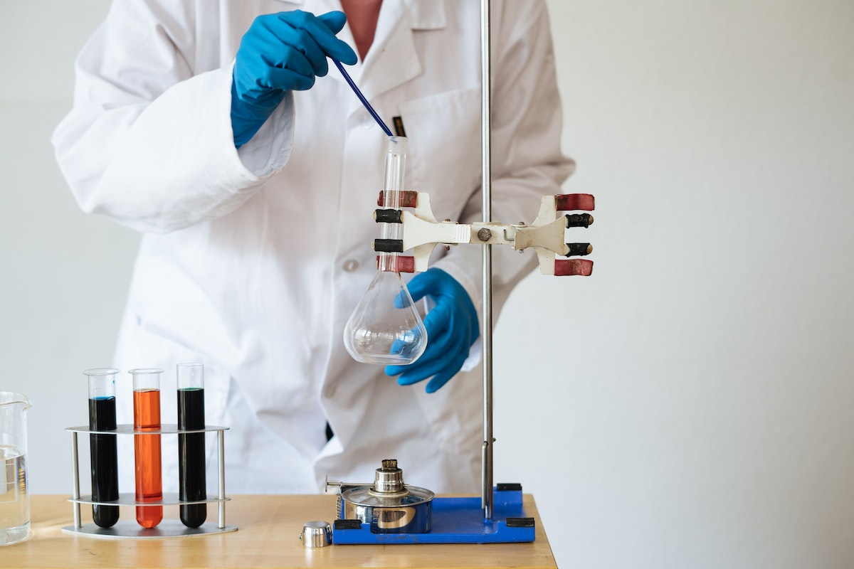 Um químico mexendo em um frasco dentro do sala de laboratório. Ele está de jaleco branco e luvas de borracha azul 