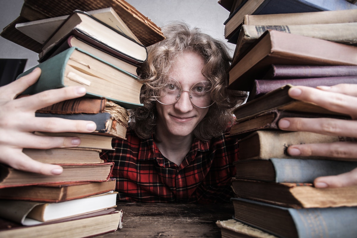 Um rapaz com óculos e cabelos longos. Ele abraça duas pilhas de livros em cima da mesa