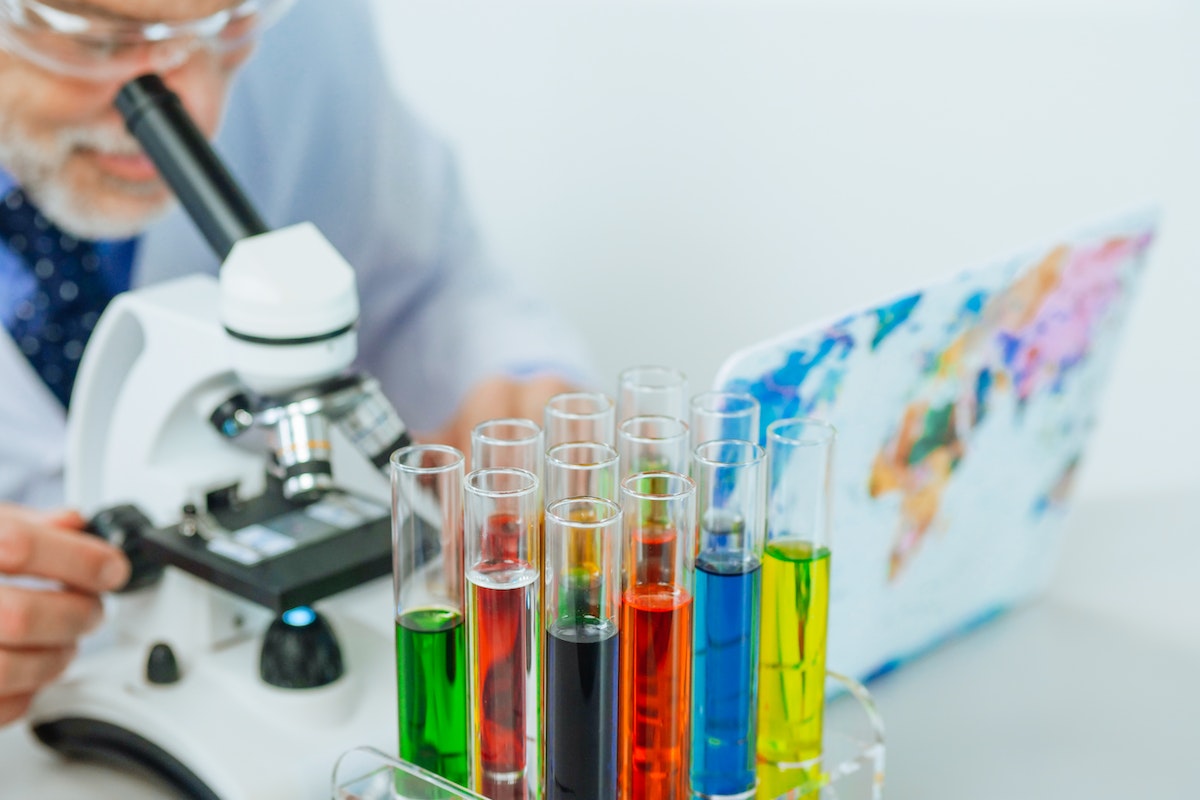 Um químico no laboratório olhando em um microscópio. Na imagem aparece diversos frascos de vidro com líquidos de cores diferentes.