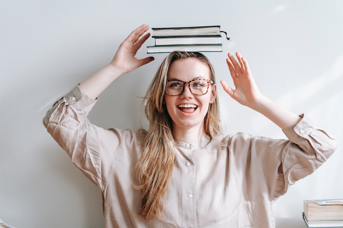 Uma estudante do Vestibular Cederj 2024 equilibrando alguns cadernos na cabeça. Ela usa óculos e está feliz sorrindo