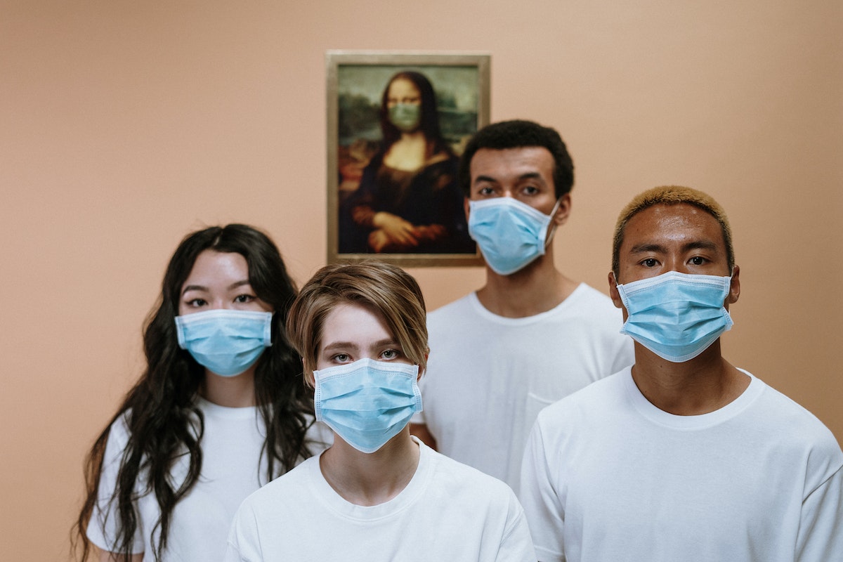 Quatro estudantes de biomedicina olhando para câmera. Eles estão de camisa branca e máscaras azul. Ao fundo, há uma parede com o quadro da Monalisa