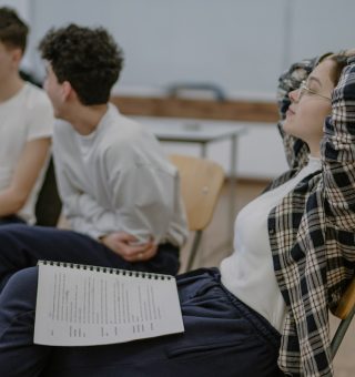 Foco em uma aluno sentada na sala de aula com a cabeça apoiada nas mãos. Ao fundo tem dois alunos