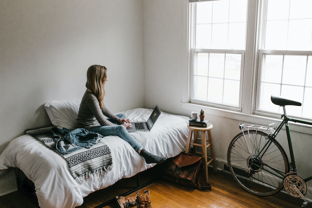 Uma mulher em seu quarto. Ela está sentada em cima da cama, mexendo em seu notebook. O quarto tem as paredes brancas, piso de madeira e uma bicicleta encostada na parede