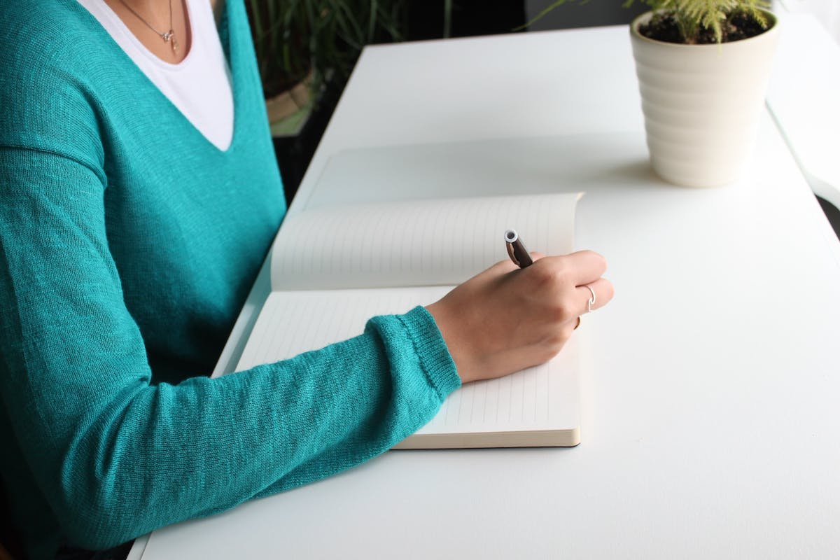 Imagem de uma mulher escrevendo em um caderno em cima de uma mesa.