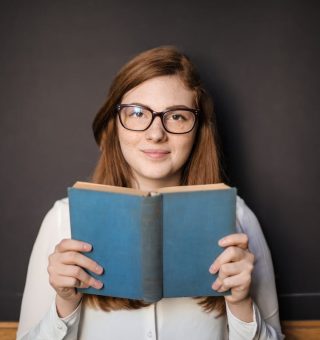 Uma menina está segurando um livro de capa azul enquanto olha e sorri para câmera. Ela está usando óculos e está na frente de um quadro negro