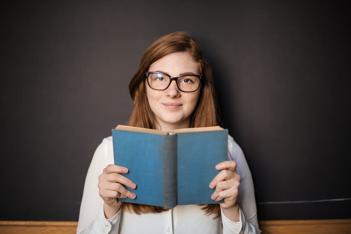 Uma menina está segurando um livro de capa azul enquanto olha e sorri para câmera. Ela está usando óculos e está na frente de um quadro negro