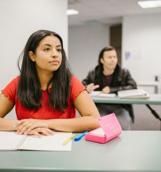 Uma aluna que se prepara para o Vestibular Unit 2024. Ela está sentada na carteira com alguns materiais. Atrás, desfocado, tem um aluno sentado em outra mesa