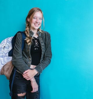 Uma menina que está Estudando para o Vestibular 2024 na frente de uma parede azul. A menina segura a mochila nas costas com uma alça e sorri