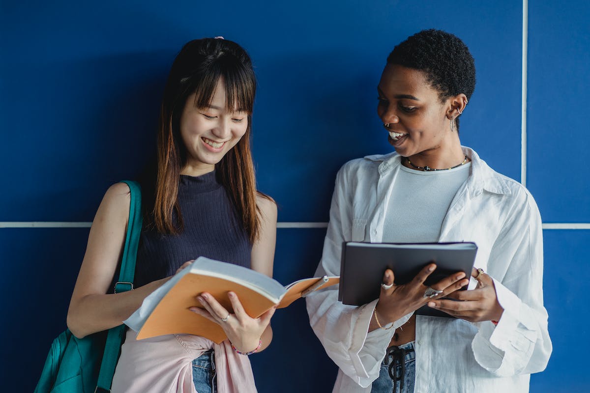 Duas amigas estão com os materiais da escola na mão. Elas sorriem e estão de pé em frente a um fundo azul