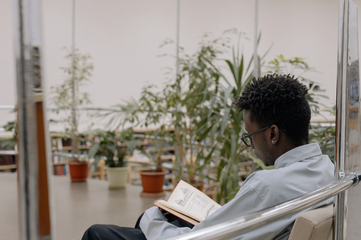 Um aluno está lendo um livro sentado, ao fundo há diversas plantas 