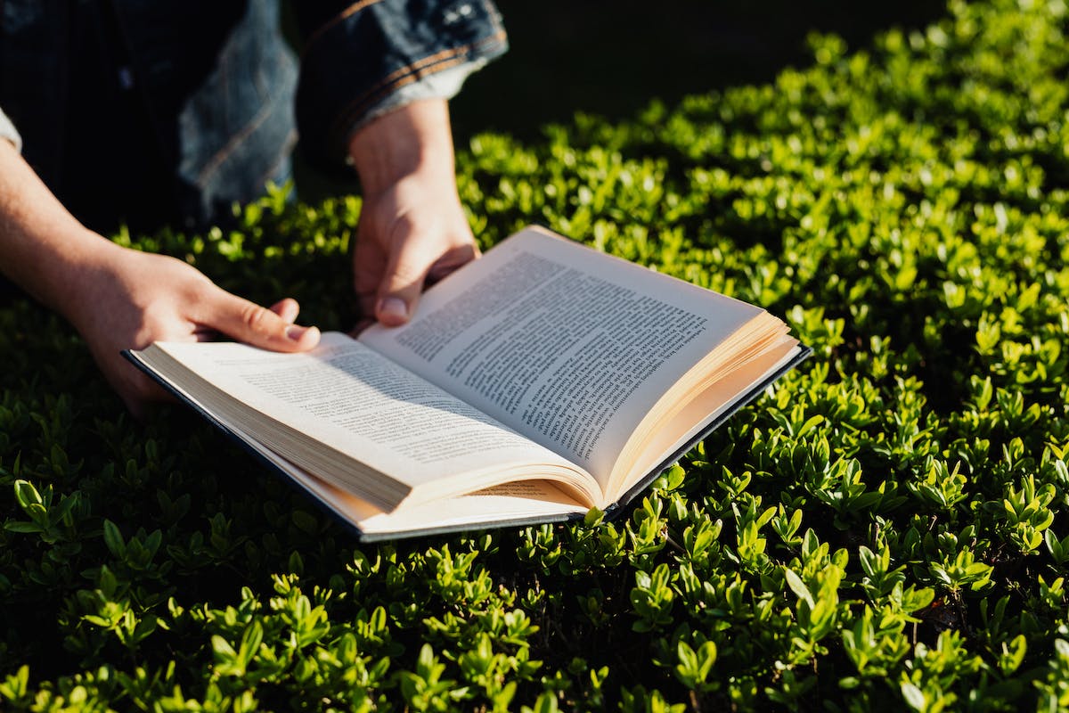 Um homem lê um livro indicado para Vestibular de Inverno 2024. Aparecem apenas suas mãos, o livro, e a grama do chão onde está sentado