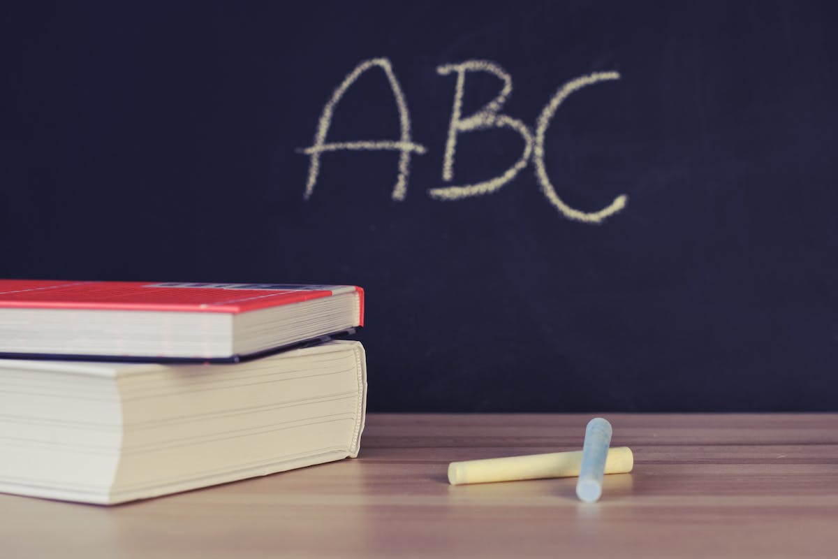 Imagem de um quadro negro onde está escrito "ABC". Dá para enxergar um giz e dois livros em uma mesa de madeira 