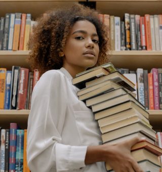 Uma menina está dentro da biblioteca e segura uma pilha de livros