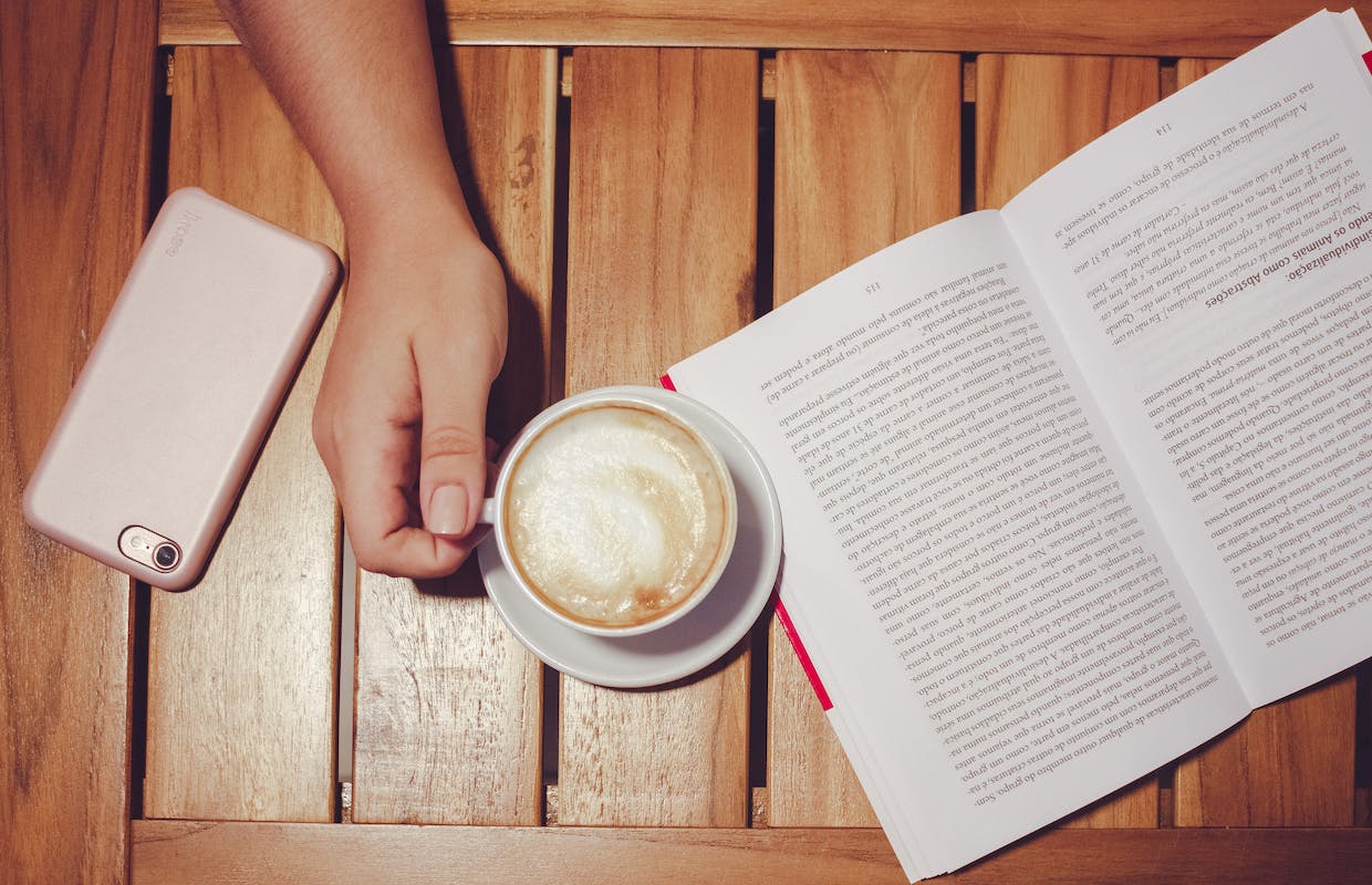 Em cima de uma mesa há o livro de um estudante e uma xícara de café com leite