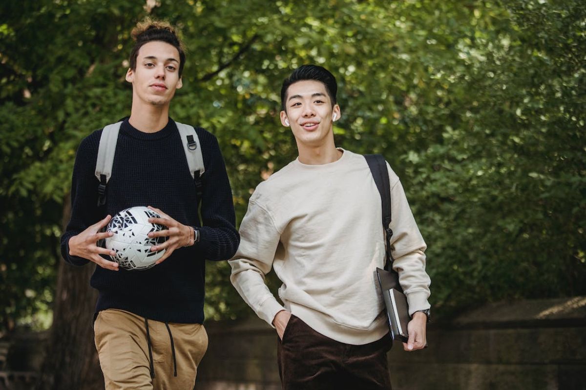 Dois alunos estão dentro de um campus. Um dele segura uma bola de futebol enquanto o outro leva uma pasta