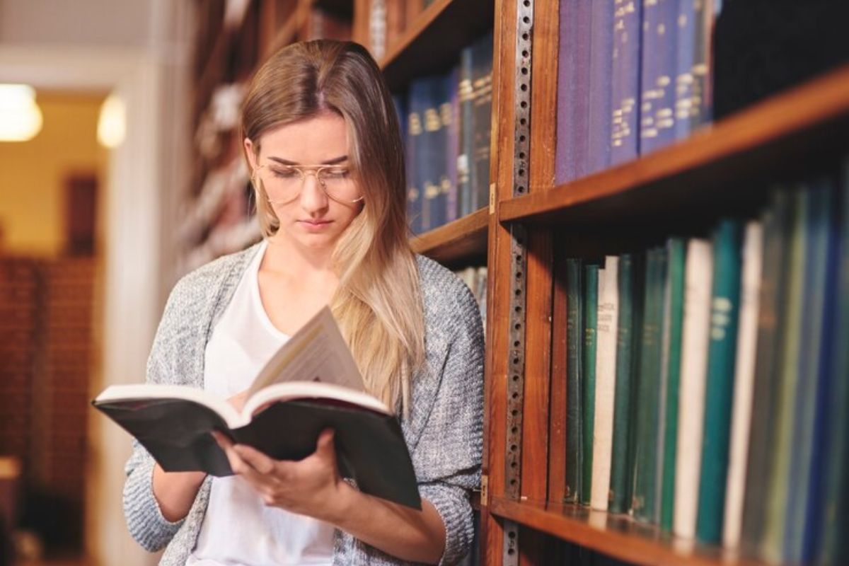 Uma aluna lendo um livro ao lado de uma estante da biblioteca