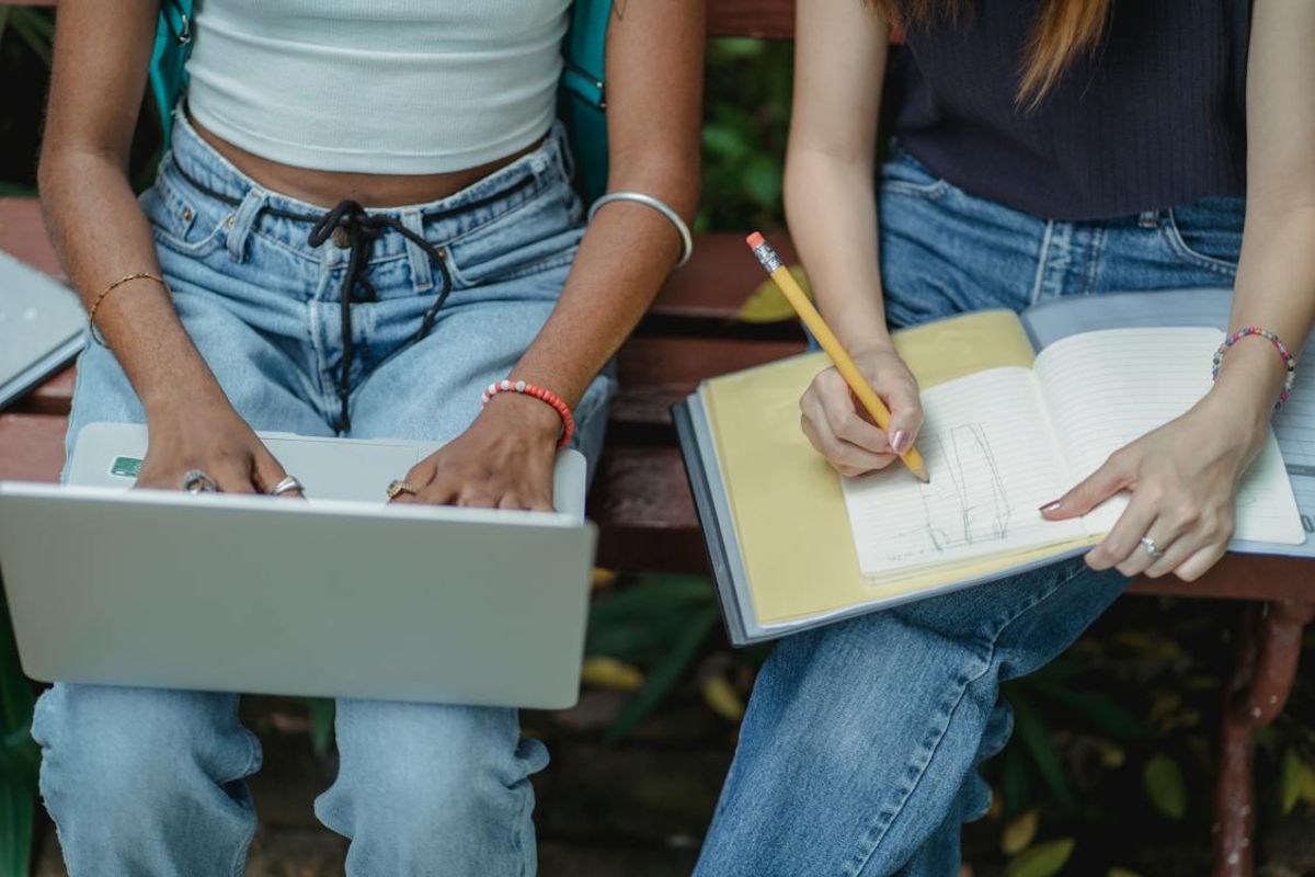Duas alunas vistas de cima enquanto estudam. Uma delas faz anotações no caderno enquanto a outra usa um notebook