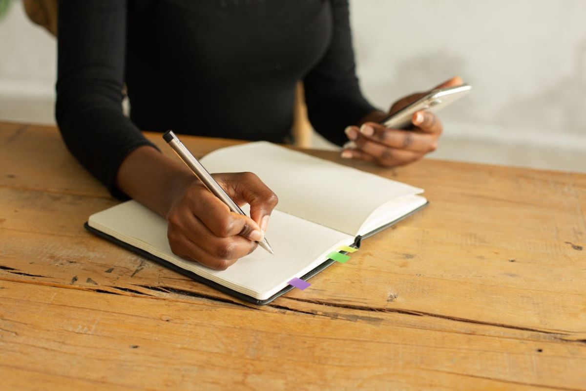A Aluna está apoiada em uma mesa enquanto escreve em um caderno com uma mão, e usa o celular com a outra