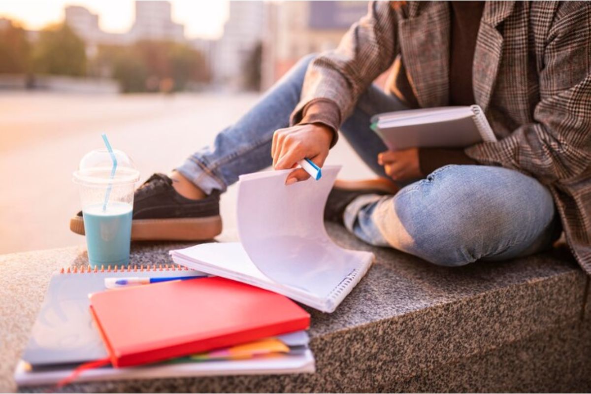 Uma estudante está sentada na mureta enquanto lê algumas páginas e estuda