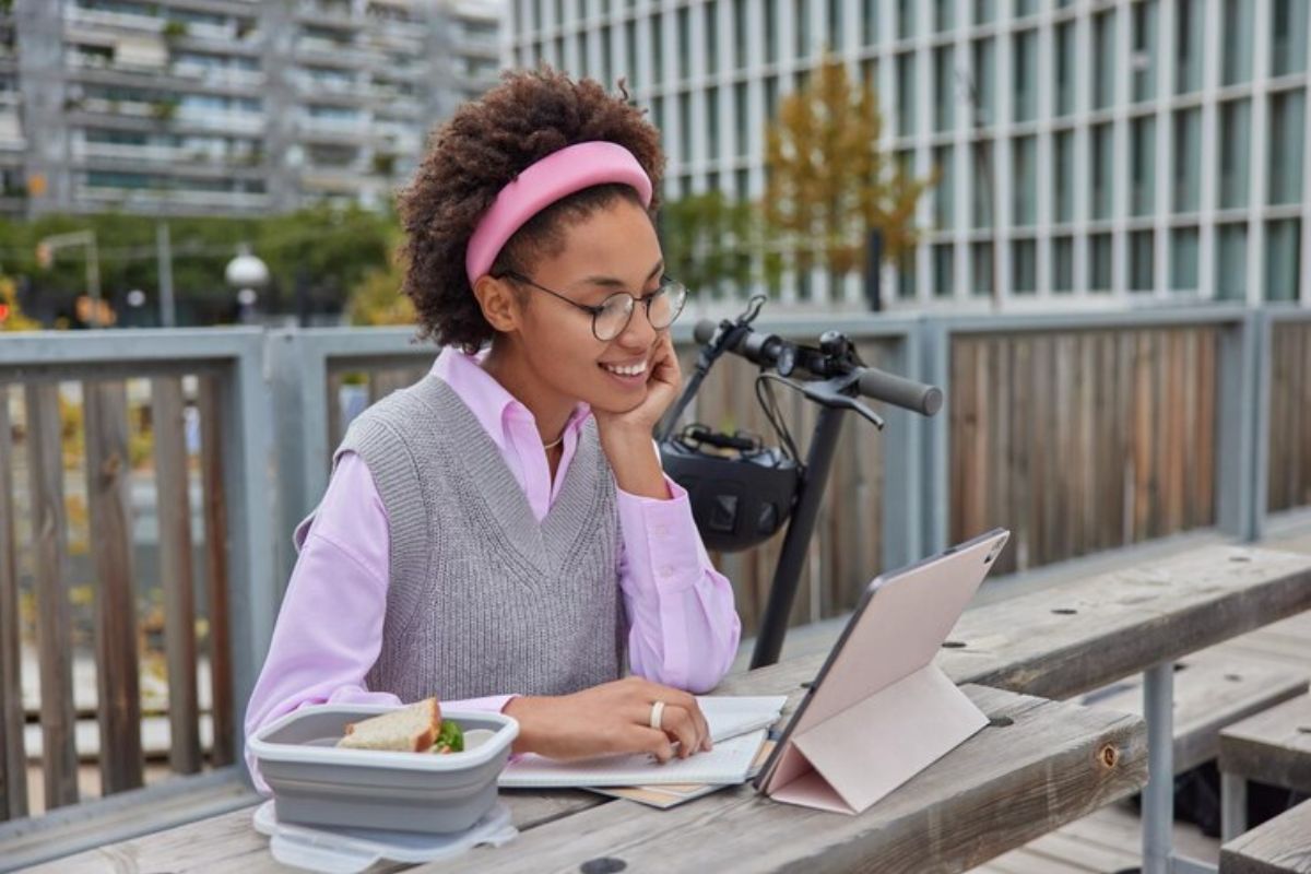 Uma aluno estuda ao ar livre. Seu tablet está em u suporte na mesa enquanto ela faz anotações no caderno